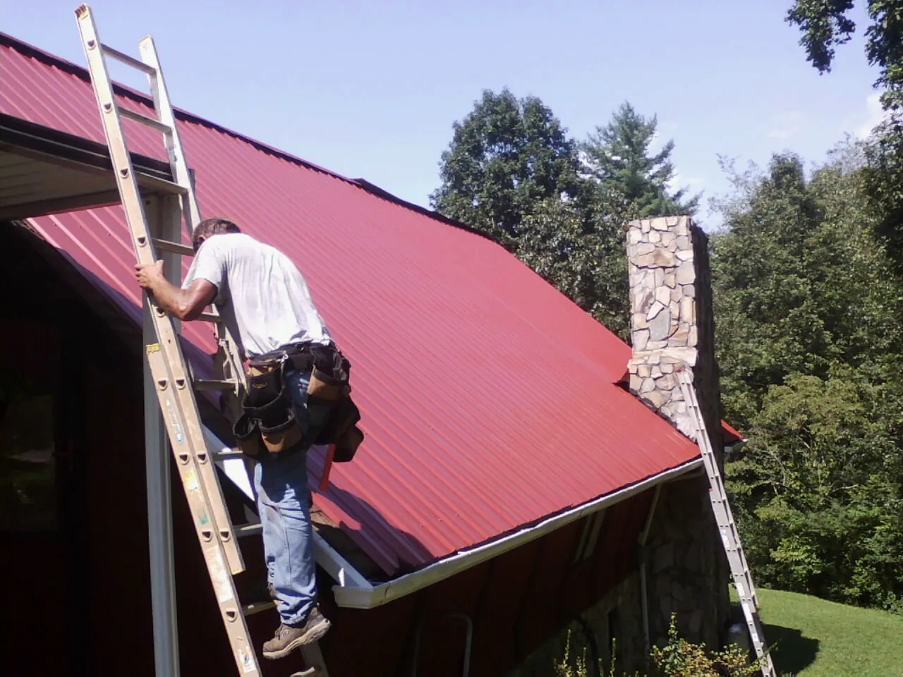 Roofer on a ladder beside a red metal roof and stone chimney — Soffit & Fascia Repair by Roofing Brothers in Sulphur Springs